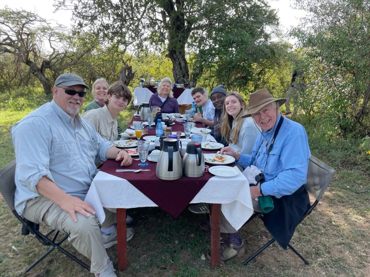 A group of people enjoying an outdoor meal together in nature surrounded by woodland.