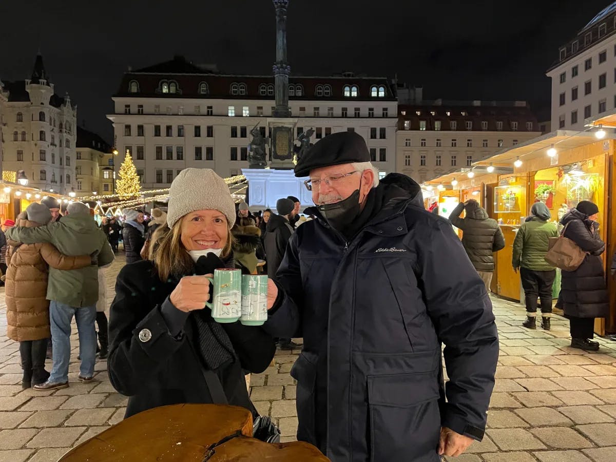 A festive Christmas market scene at night with two people in winter coats holding drinks and enjoying the atmosphere