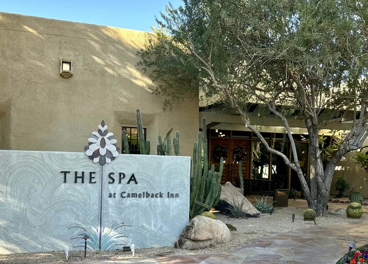 A stone wall that reads "Spa at Camelback Inn" outside of a desert-colored building with a tree and cactus outside.