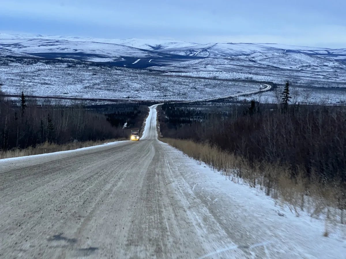 An icy highway with snow covered mountains in the distance and pine trees on the right and left side of the road.