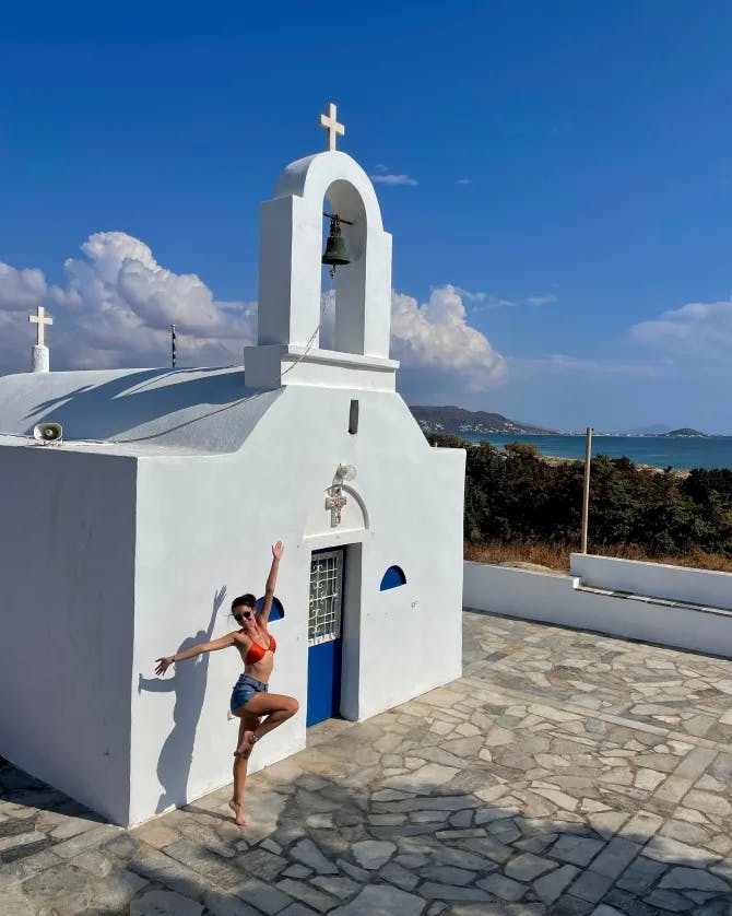 Alayna with her arms in the air standing on one foot in front of a small white building under sunny skies