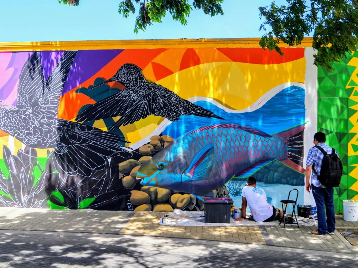 A colorful street mural with artistic elements and people interacting, under a clear blue sky.