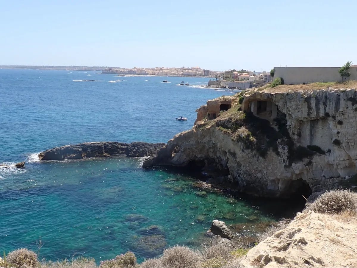 The image shows a coastal seascape with clear blue waters, a rocky cliffside, and buildings in the distance under a bright sky.