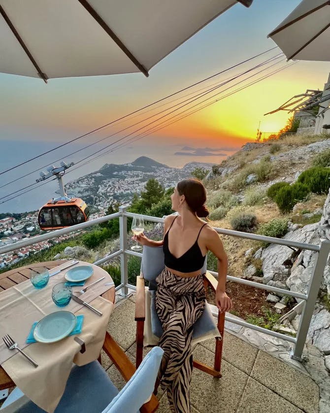 Camille seated at a dining table outdoors on a balcony overlooking a coastal area at sunset