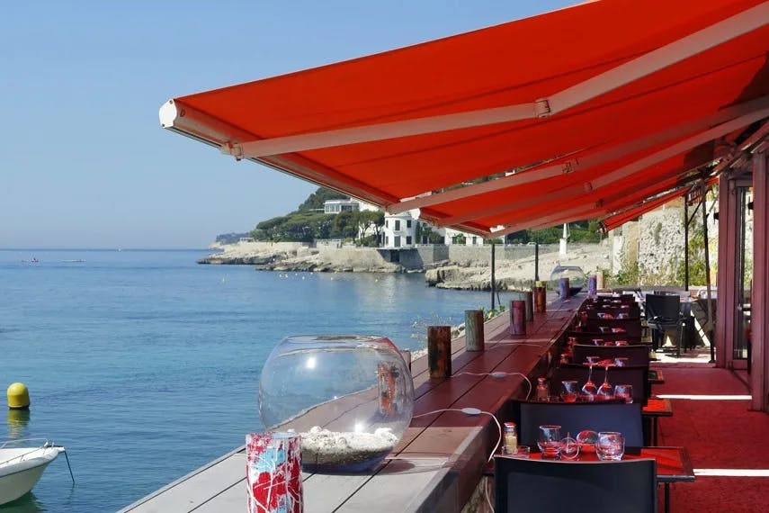 An outdoor restaurant seating area next to the water with a red canopy above