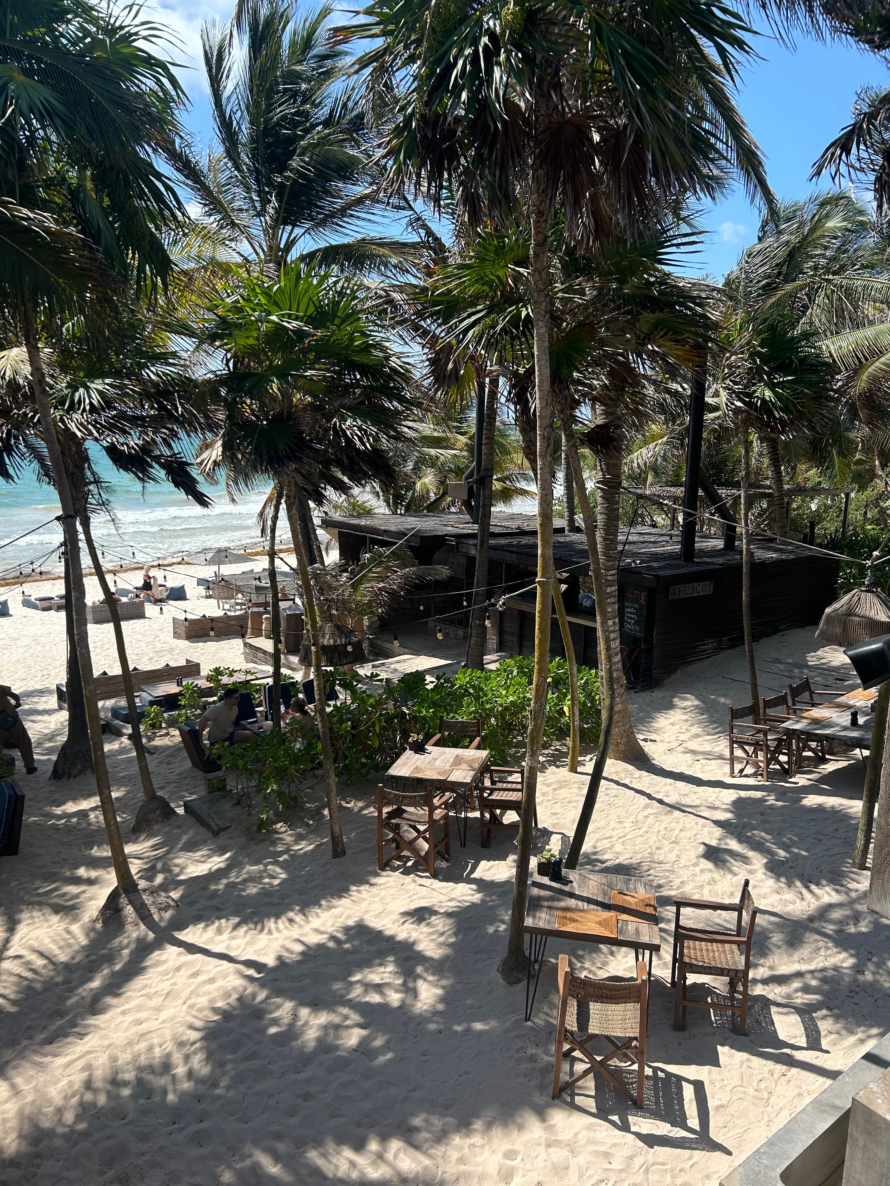 A view of the beach at Be Tulum hotel with wooden chairs and tables and palm trees