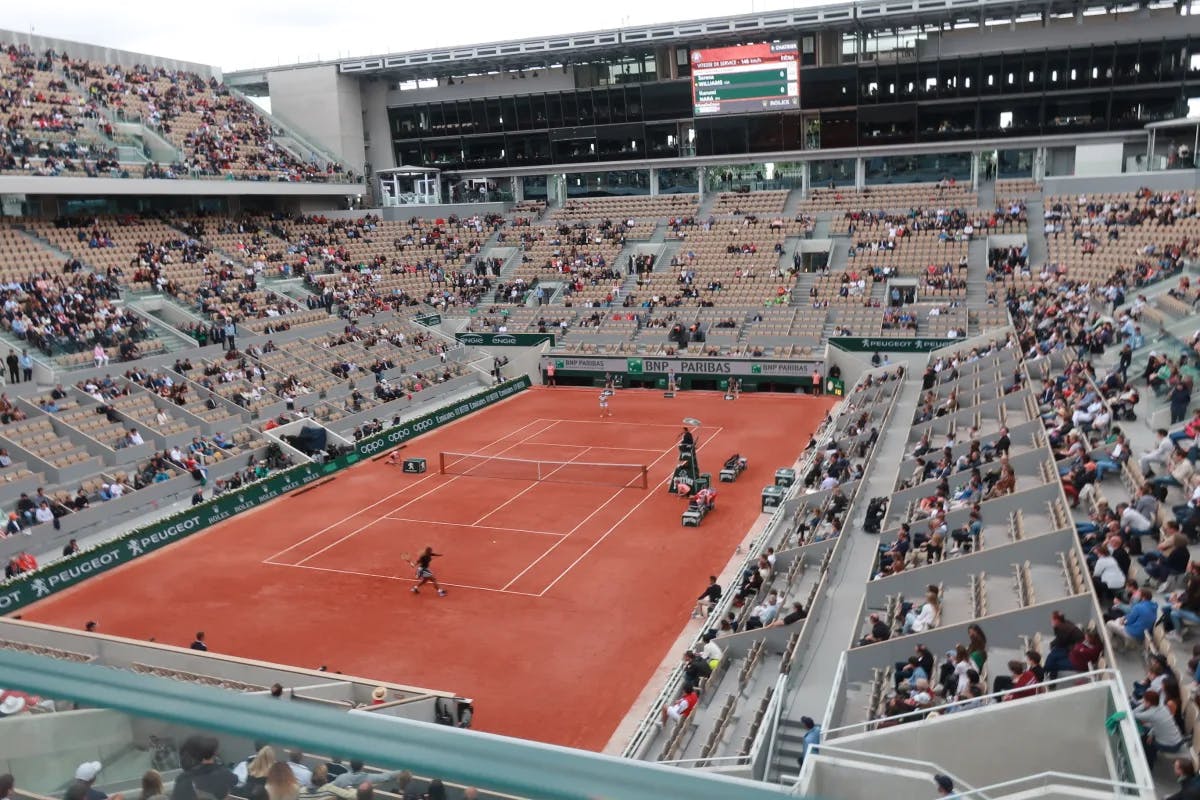 An overhead view of a red tennis court with people standing on the court, and stands scattered with various fans and viewers. 