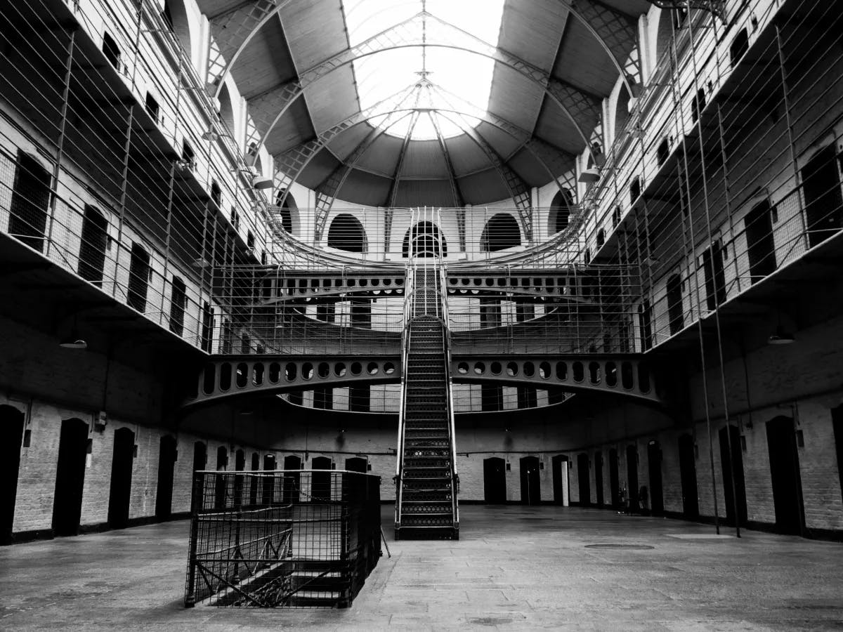 The image depicts the interior of a large, multi-level prison hall with cells on each level, a central staircase, and a domed ceiling. The stark and symmetrical architecture is quite striking.