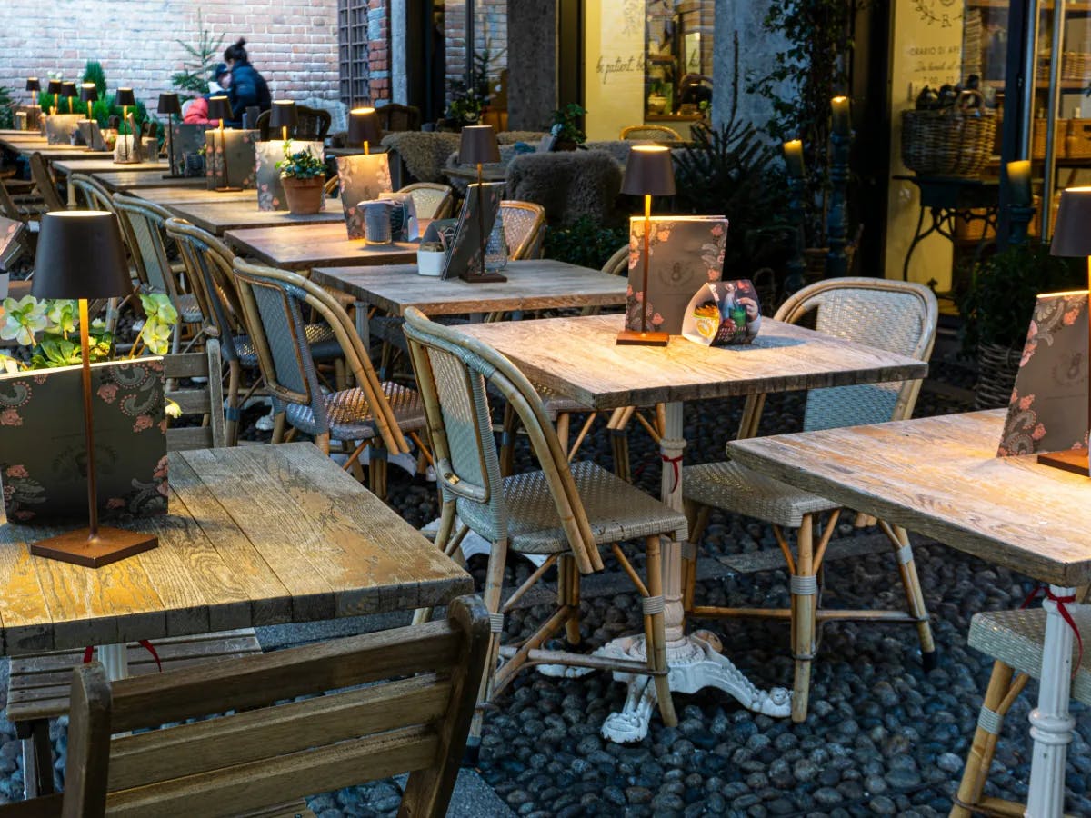 A patio with wooden tables and chairs, featuring table lamps, menus and a cobblestone flooring. 