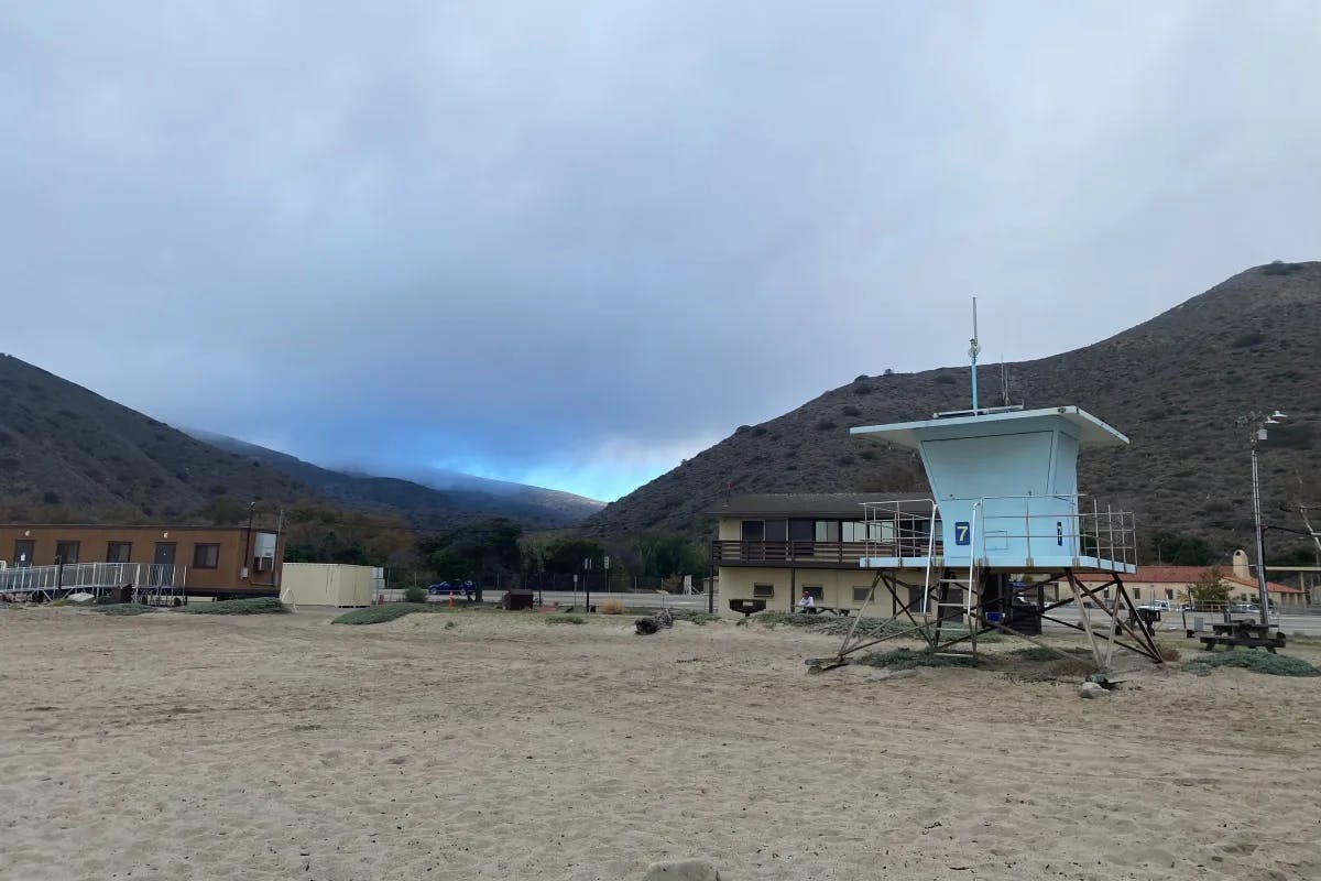 A light blue lifegraurd shack stands on a sandy beach with some other buildings behind it as well as a mountain canyon in the distance. 