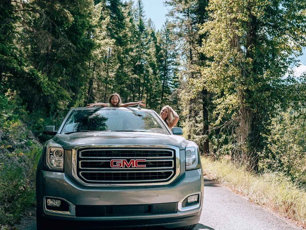 A picture of kids hanging out of the car in the woods during the daytime.