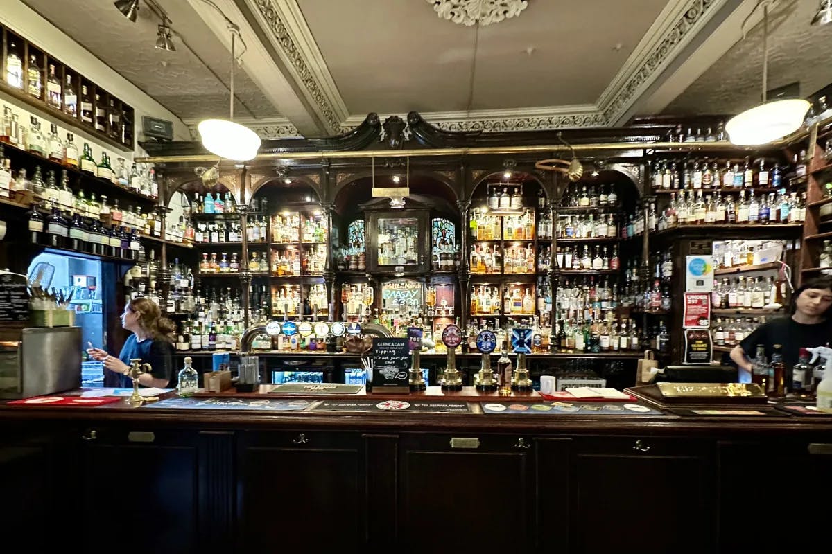 An old bar full of various bottles of alcohol, hanging lights and employees behind the wooden counter. 