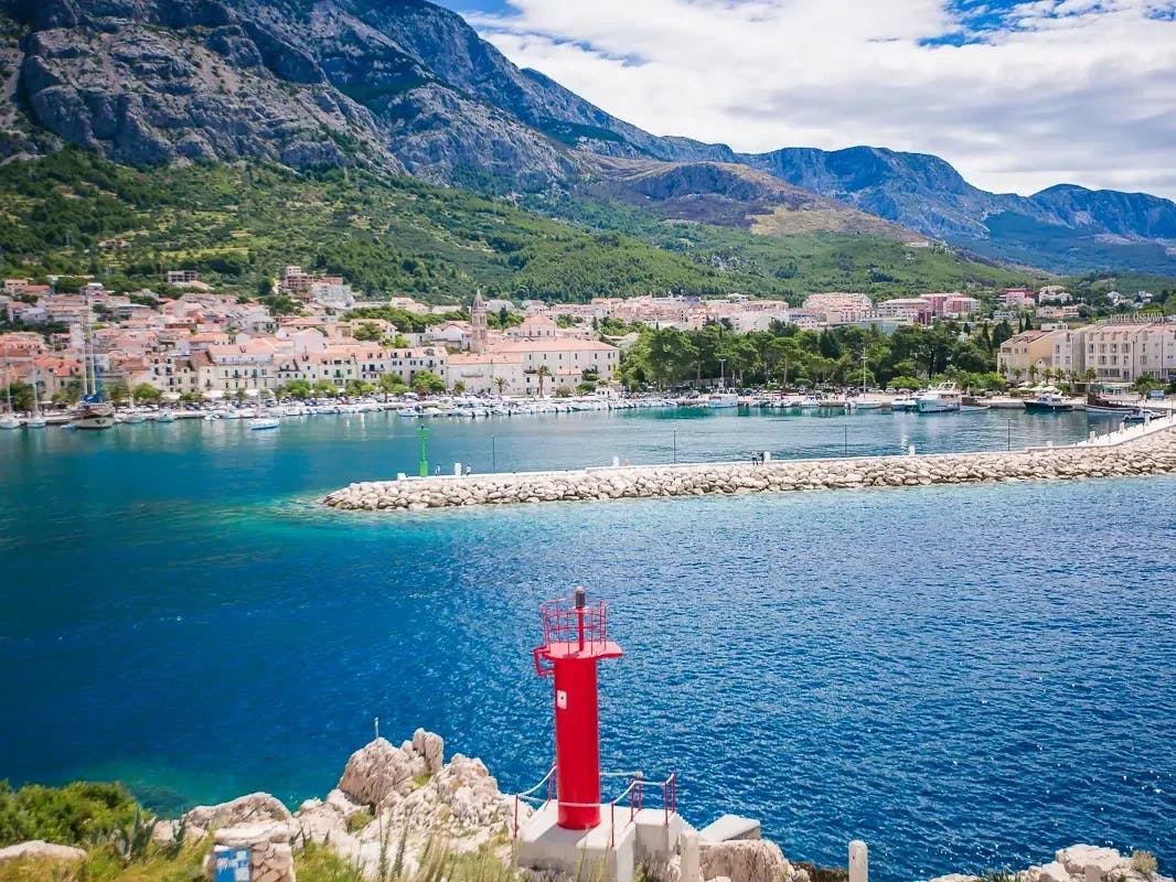 A picture of a harbor with a red piece of equipment in the forefront, and a town, trees and rocky mountain in the distance. 