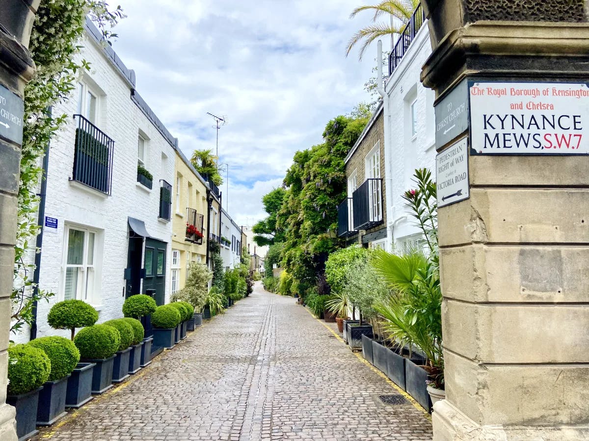 A brick road in London surrounded by buildings on each side, potted green plants, trees and a sign at the entrance. 