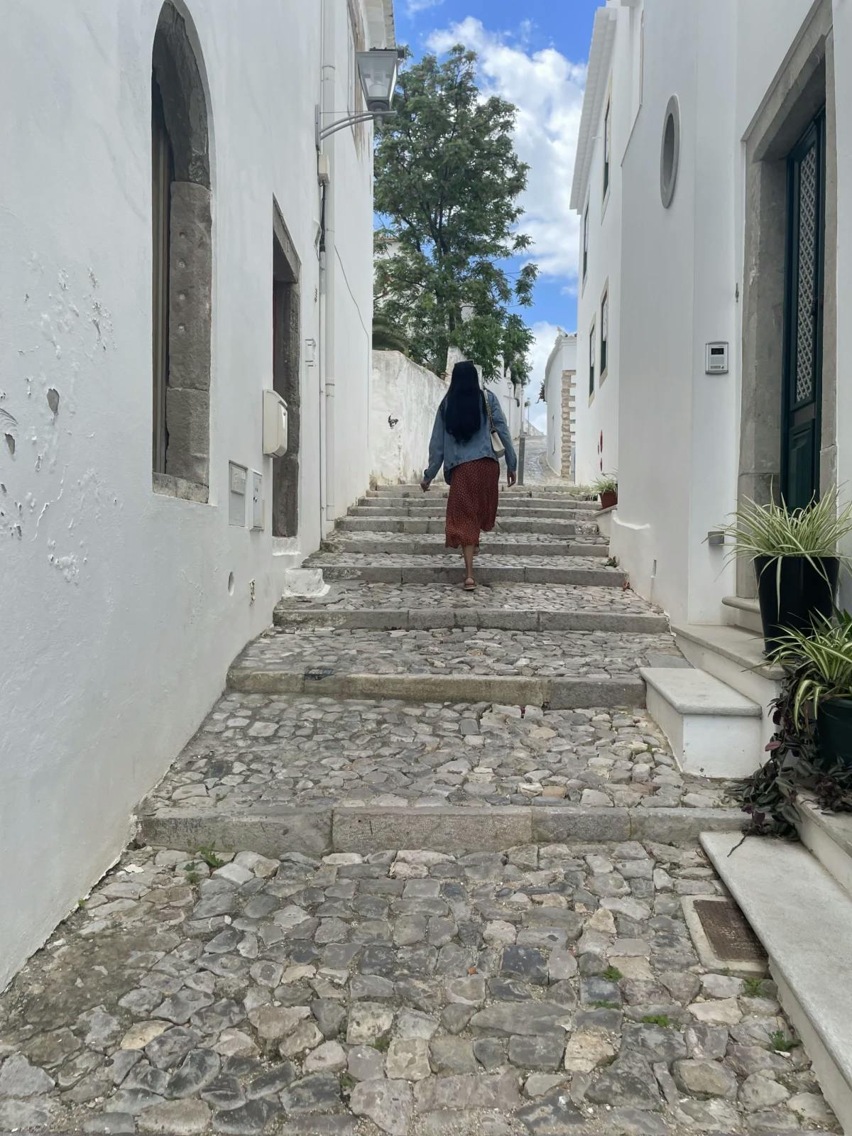 A point of view look through a narrow alleyway of Tavira, with a women walking away from the camera, up the stone steps, towards the sunshine. 