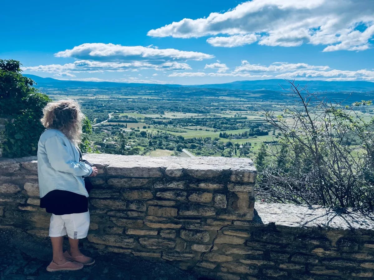 A person admiring a scenic landscape from behind a stone wall under a cloud-streaked blue sky.