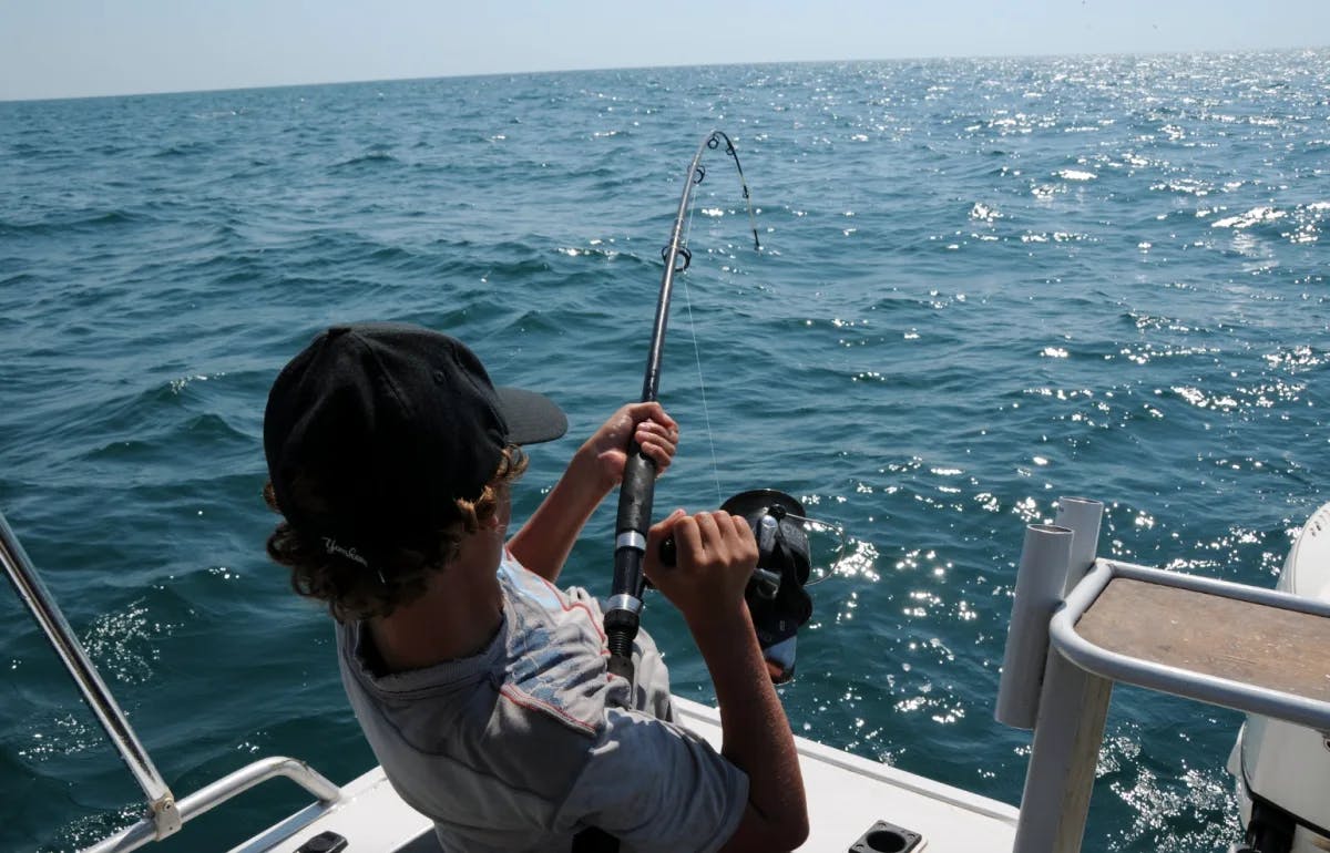 A man standing on the edge of a boat while holding a fishing rod and luring a catch in from the sparkling blue water.