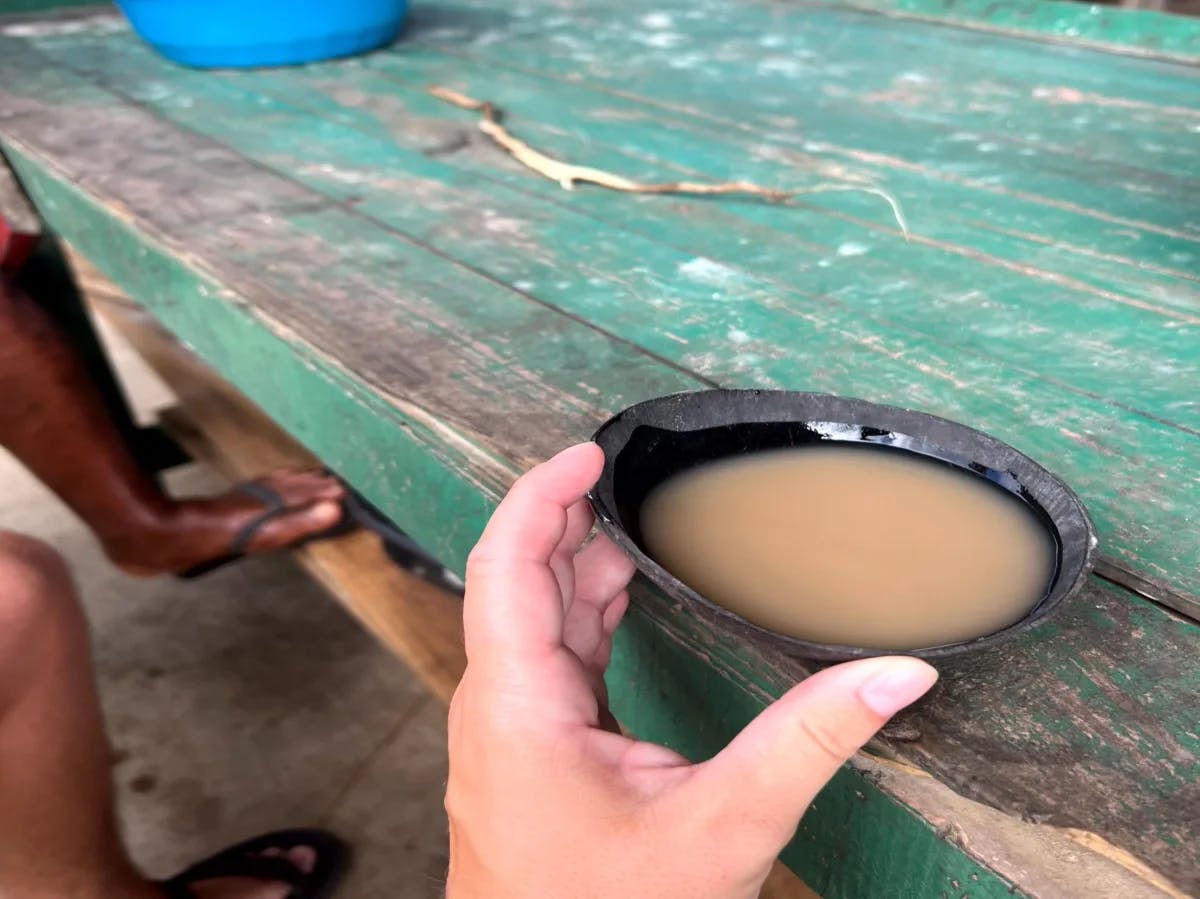 A hand reaching towards a small black bowl filled with kava on a wooden surface.