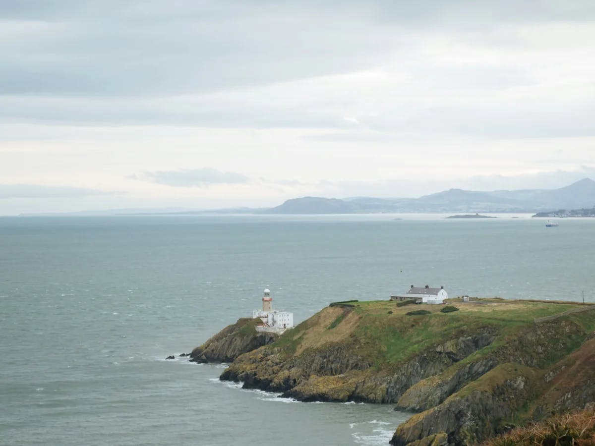A lighthouse perched on a cliff, overlooking the sea against a backdrop of mountains and cloudy skies, at Howth.