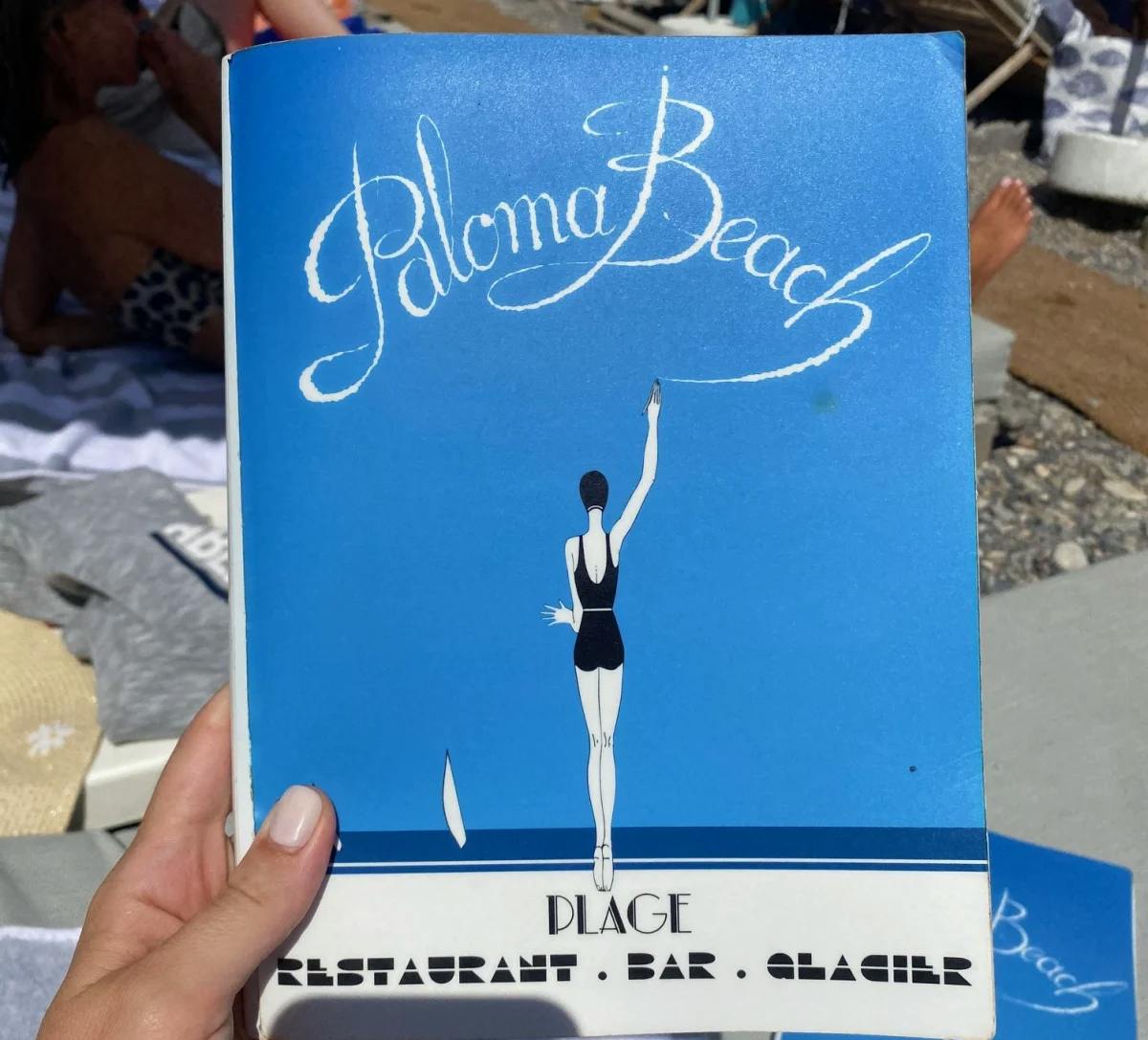 The image depicts a hand holding a book titled “Paloma Beach” with a beach scene in the background, evoking a leisurely atmosphere.