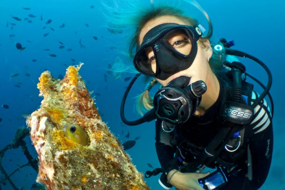 A woman scuba diving posing by coral with a fish peeping out.