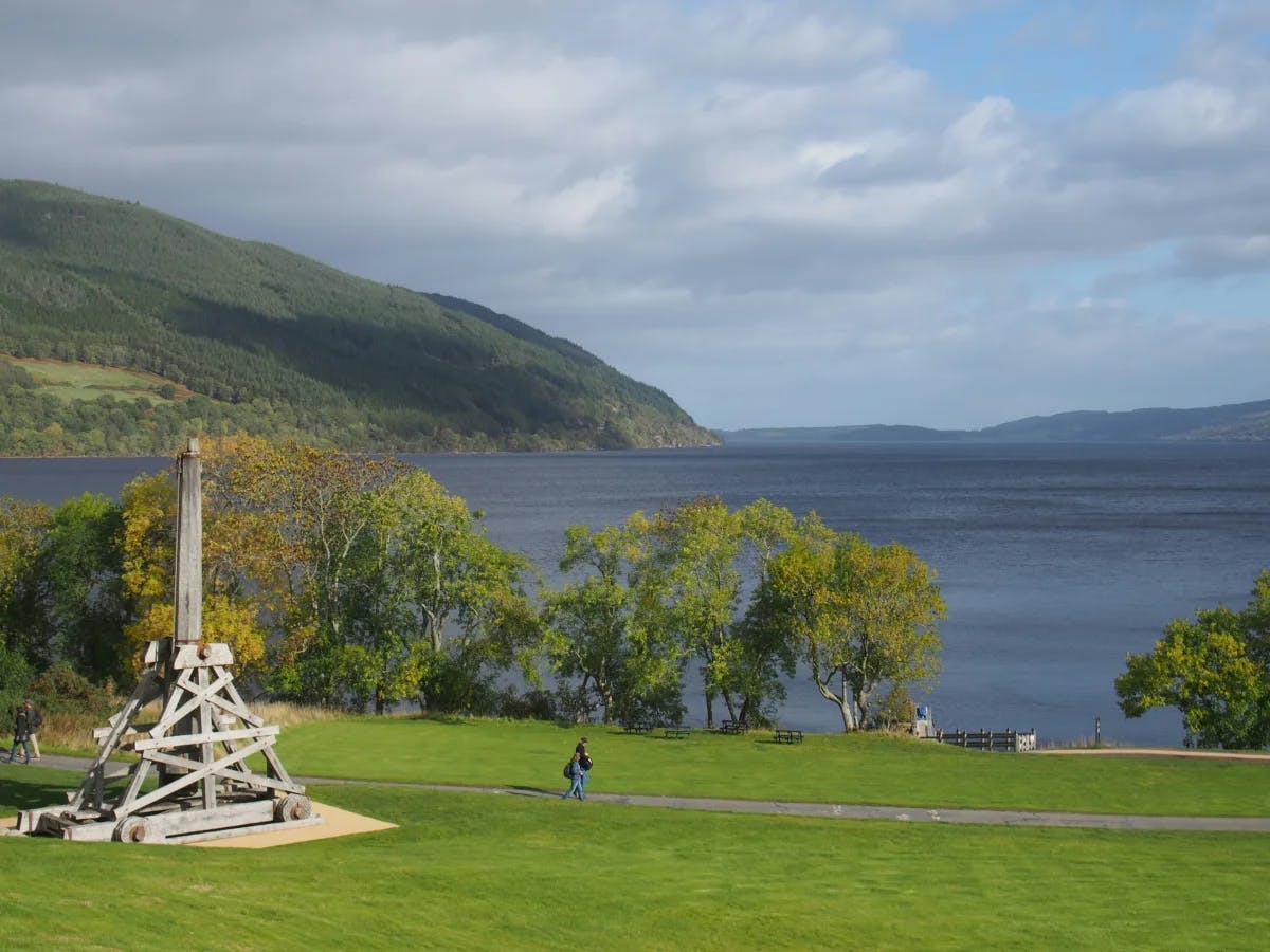 The image presents a serene lakeside view with lush greenery and a quaint wooden structure, under a clear sky with distant hills.