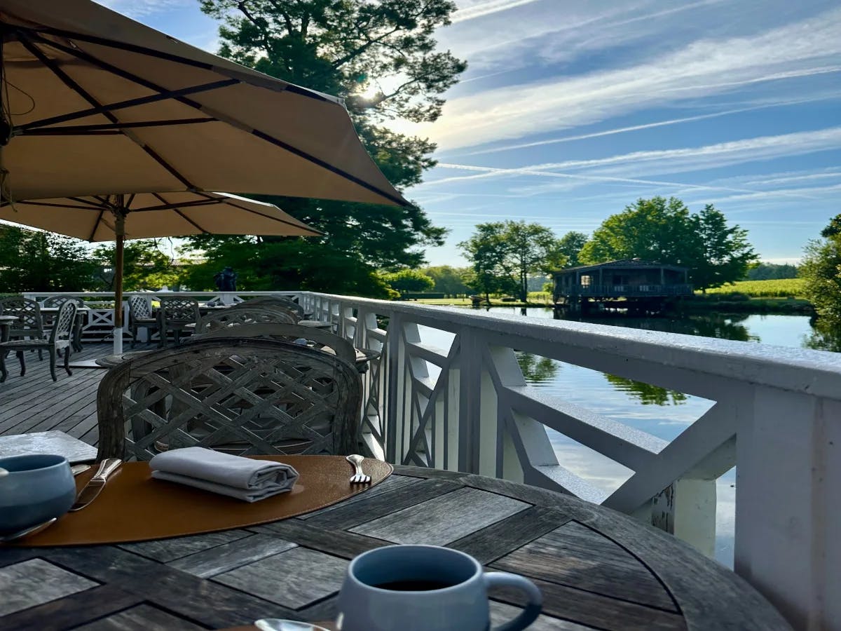 The image offers a tranquil outdoor dining experience with a table set under an umbrella, overlooking a calm body of water and verdant trees.