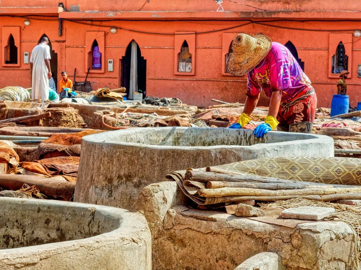 The image portrays a person engaged in traditional fabric dyeing or tanning, surrounded by vivid colors and cultural architecture.