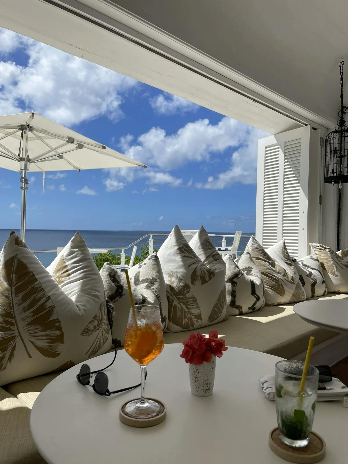 Drinks on a white table with a row of white patterned cushions behind by a large window with white shutters with ocean view