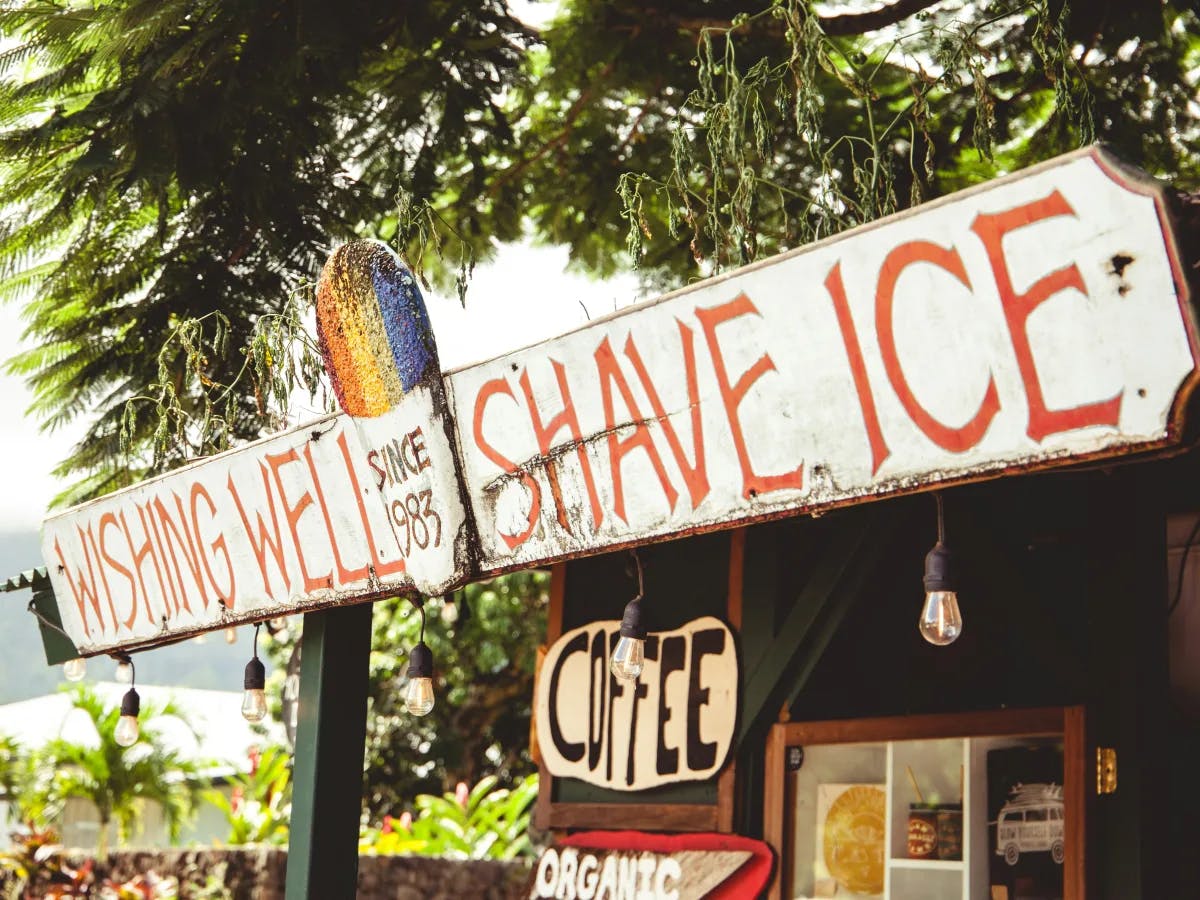 A rustic signpost with multiple directions for “Shave Ice,” “Coffee,” and “Wishing Well Shave Ice” amidst lush greenery.