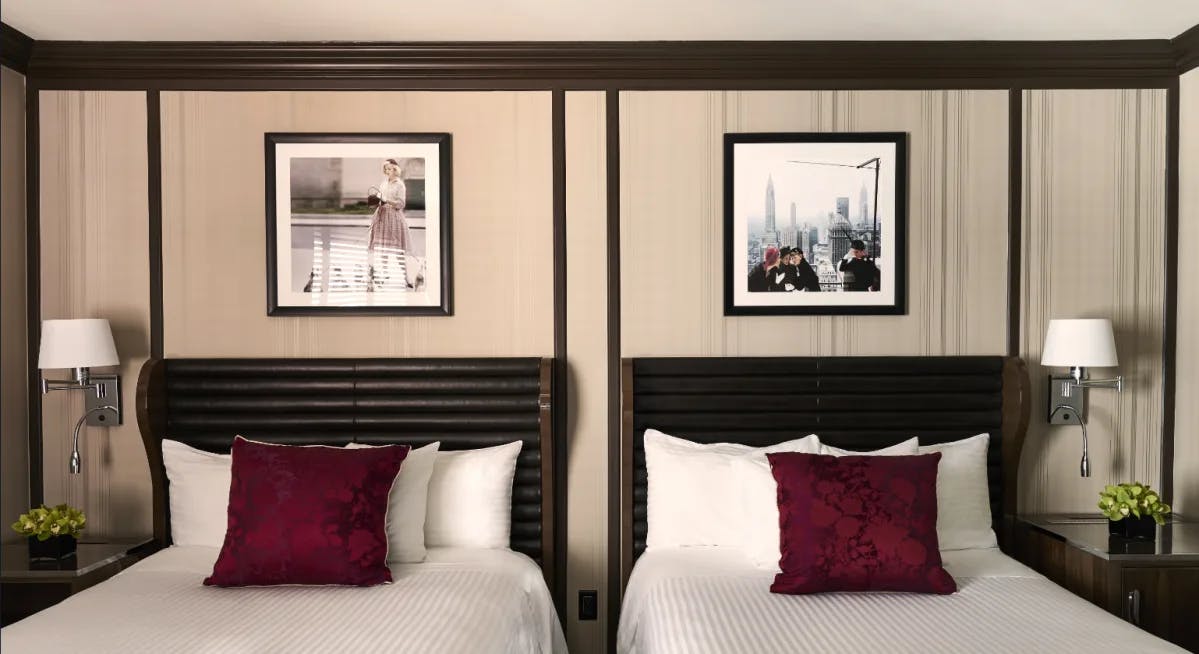 A view of a hotel room at Triumph hotels in NYC with two beds with white bedding and purple cushion and images of NYC on the wall above.