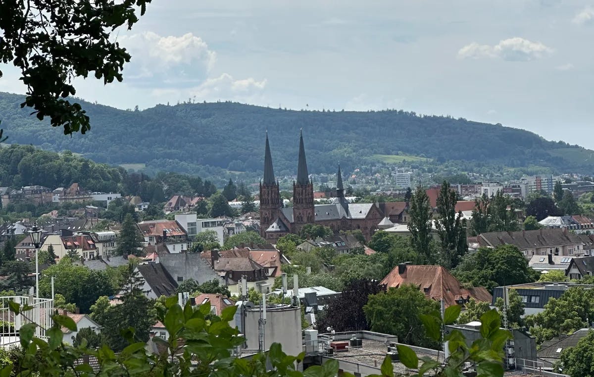 A serene village with a church tower, nestled in lush greenery against a backdrop of rolling hills and a cloudy sky.