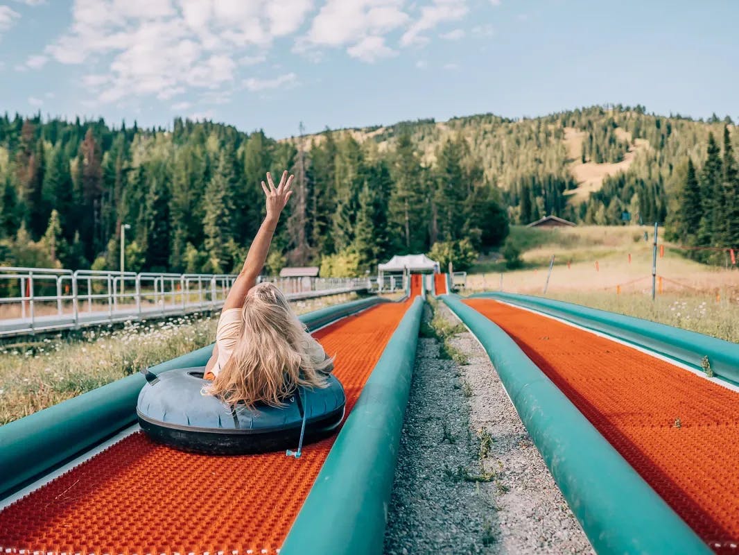 A picture of a person sliding down a hill while sitting on an inner tube. Their hand is raised above their head, and there are pine trees and a railing with stairs in the surrounding areas. 