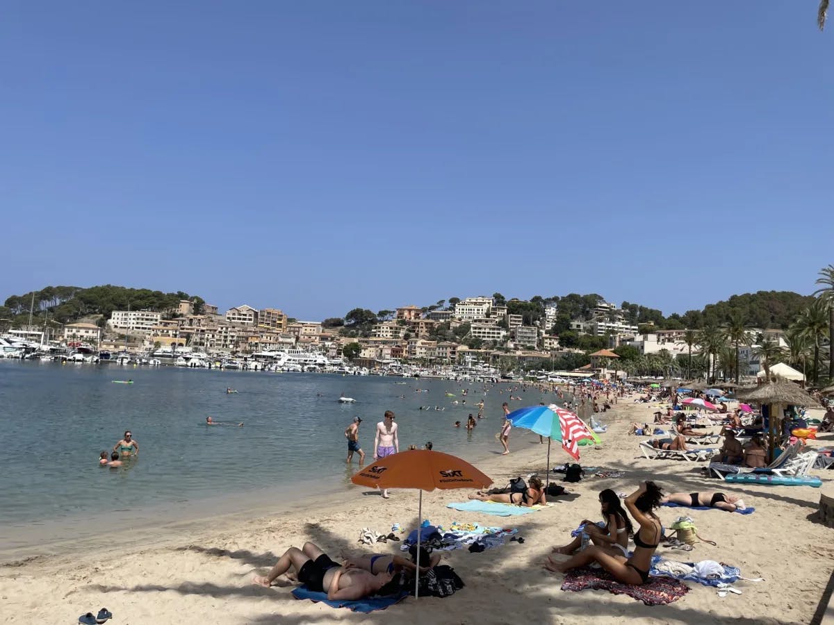 A beach scene with people laying on the sand or chairs and under umbrellas on a sunny day, with local ships and buildings in the background.