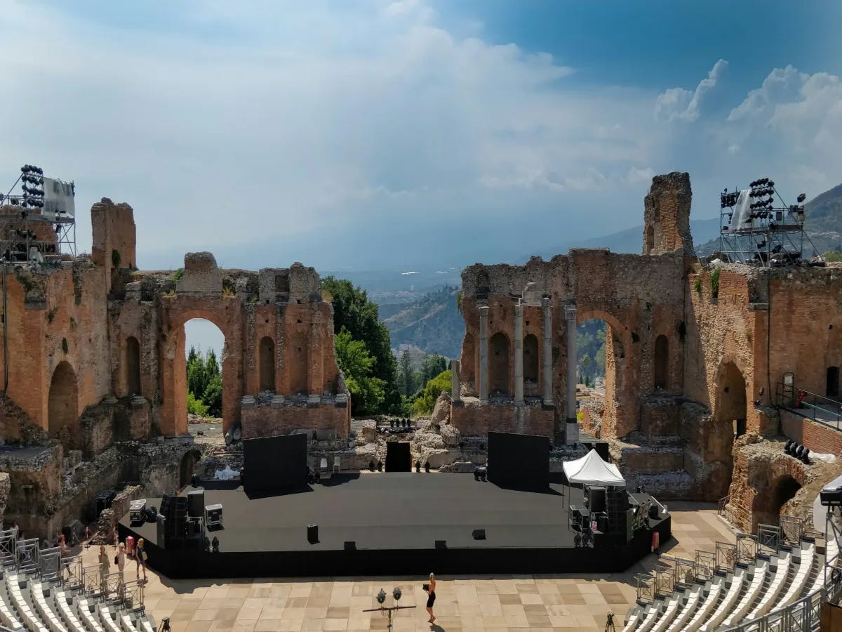 An ancient amphitheater with a modern stage setup, overlooking a scenic view of hills with greenery.