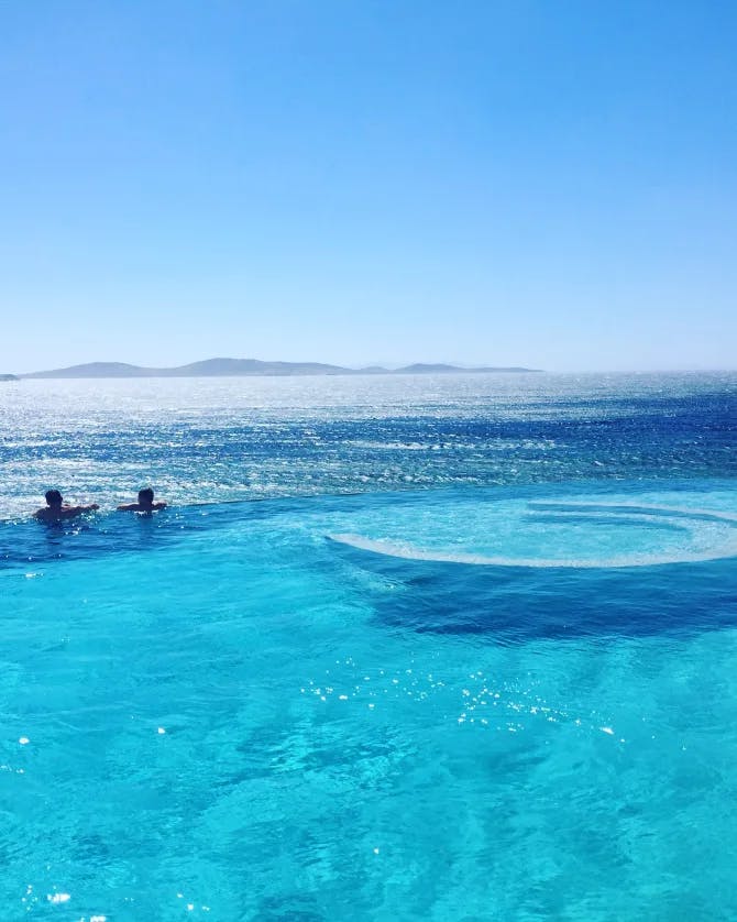 A photo of a blue pool with two people in the water overlooking the sea with the sun sparkling overhead