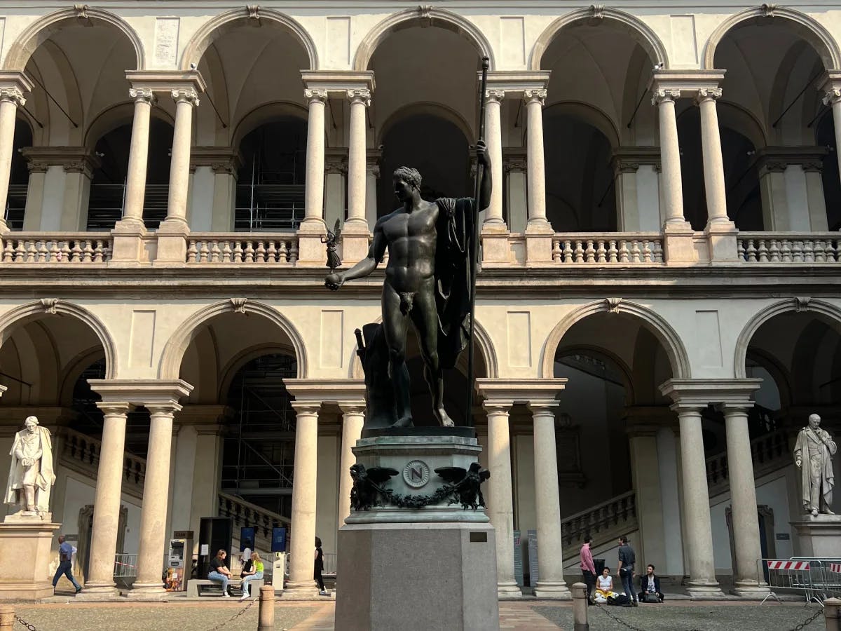 A view of a dark statue in front of arches and balconies, with two statues in the background and people walking around in a museum. 