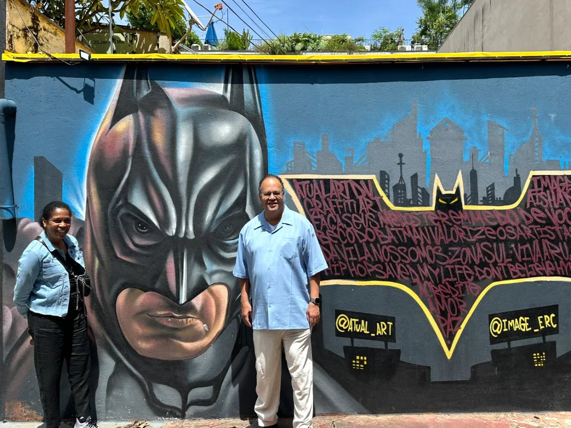 The image depicts two individuals posing in front of a vibrant Batman mural with a stylized cityscape and graffiti in the background.