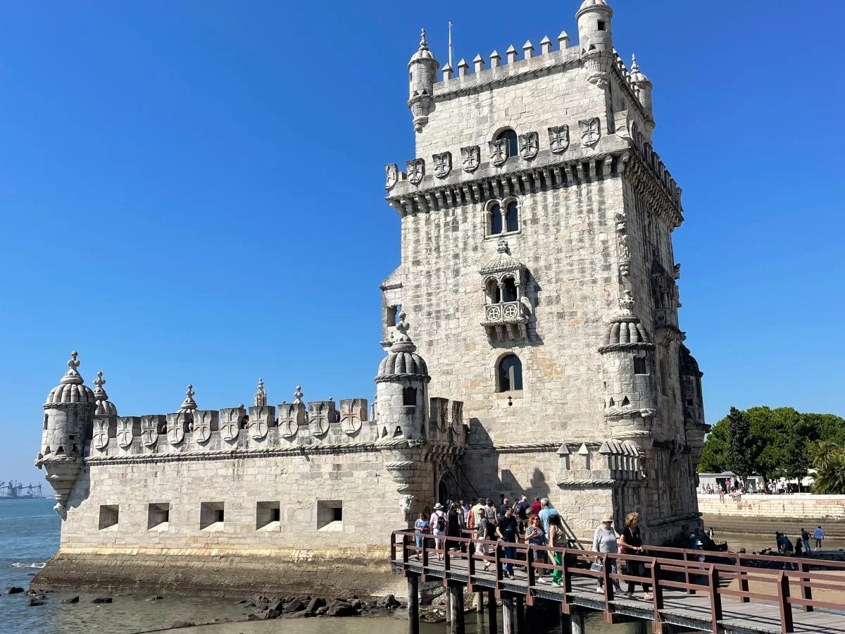 A view of the Belem tower during daytime surrounded by tourists, trees and blue water.