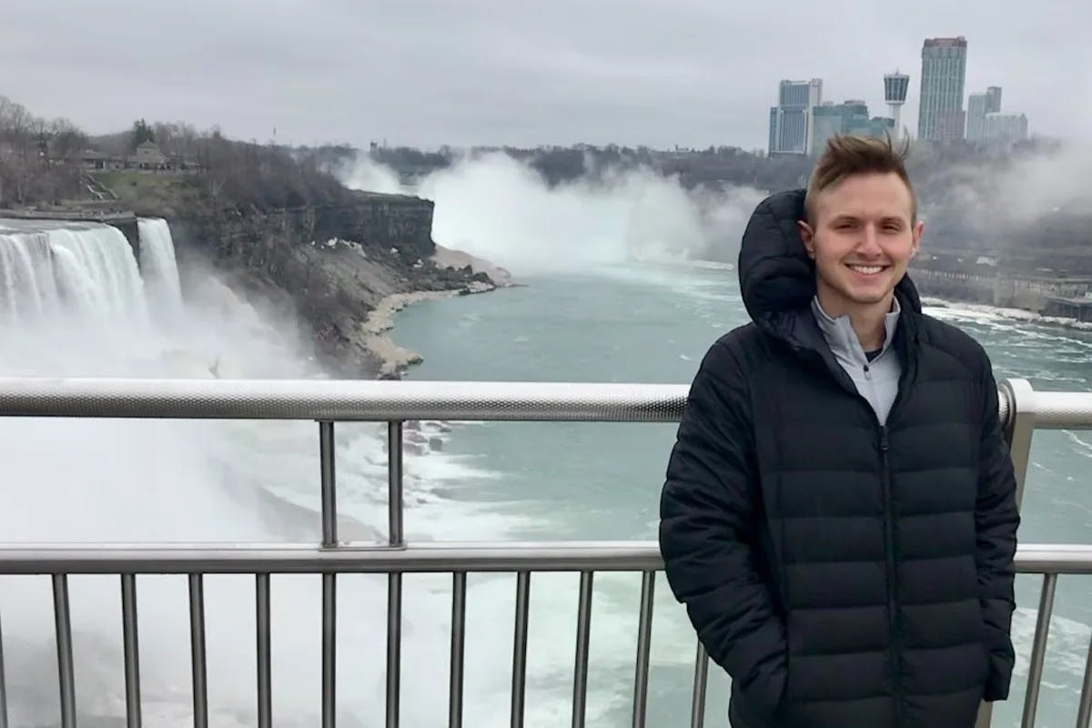 A man smiling for the camera while standing in front of a fence with the top of Niagara Falls behind it. 