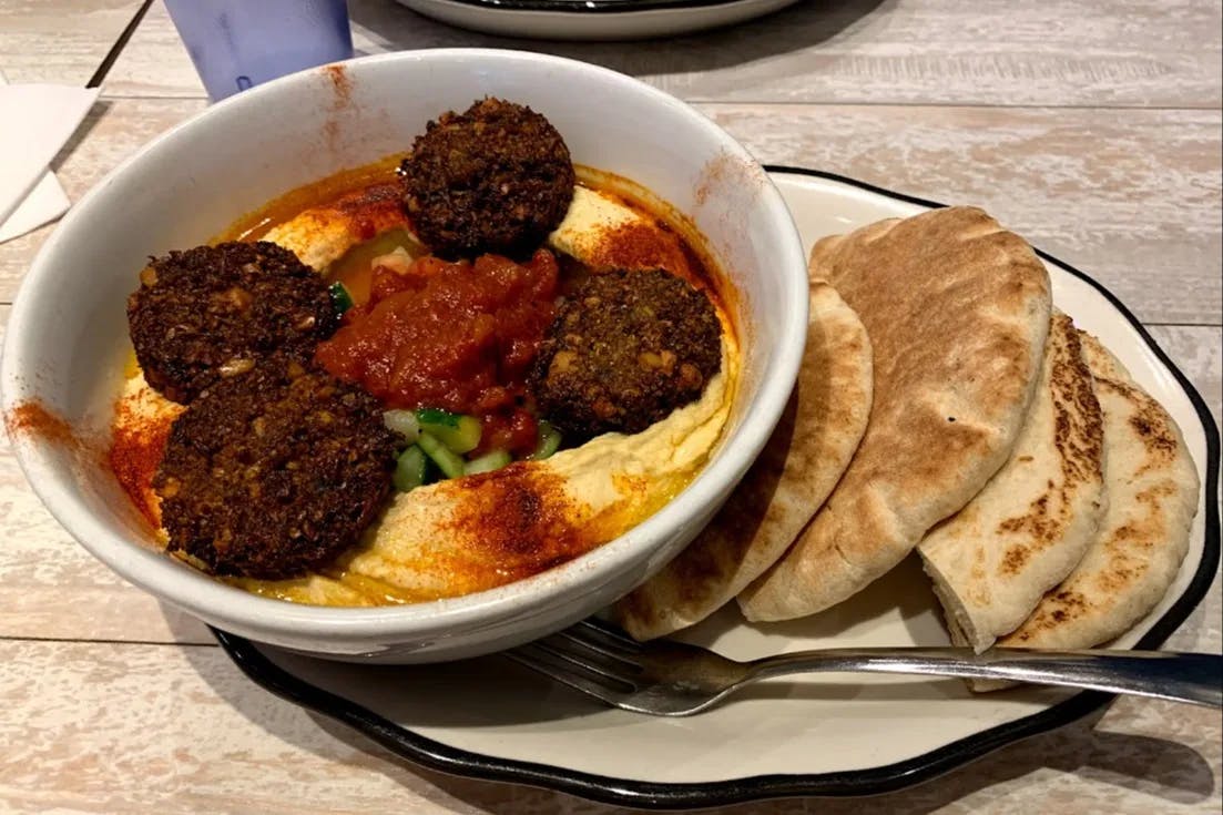 A bowl of hummus, falafel and side of pita bread on a plate sitting on a wooden table.