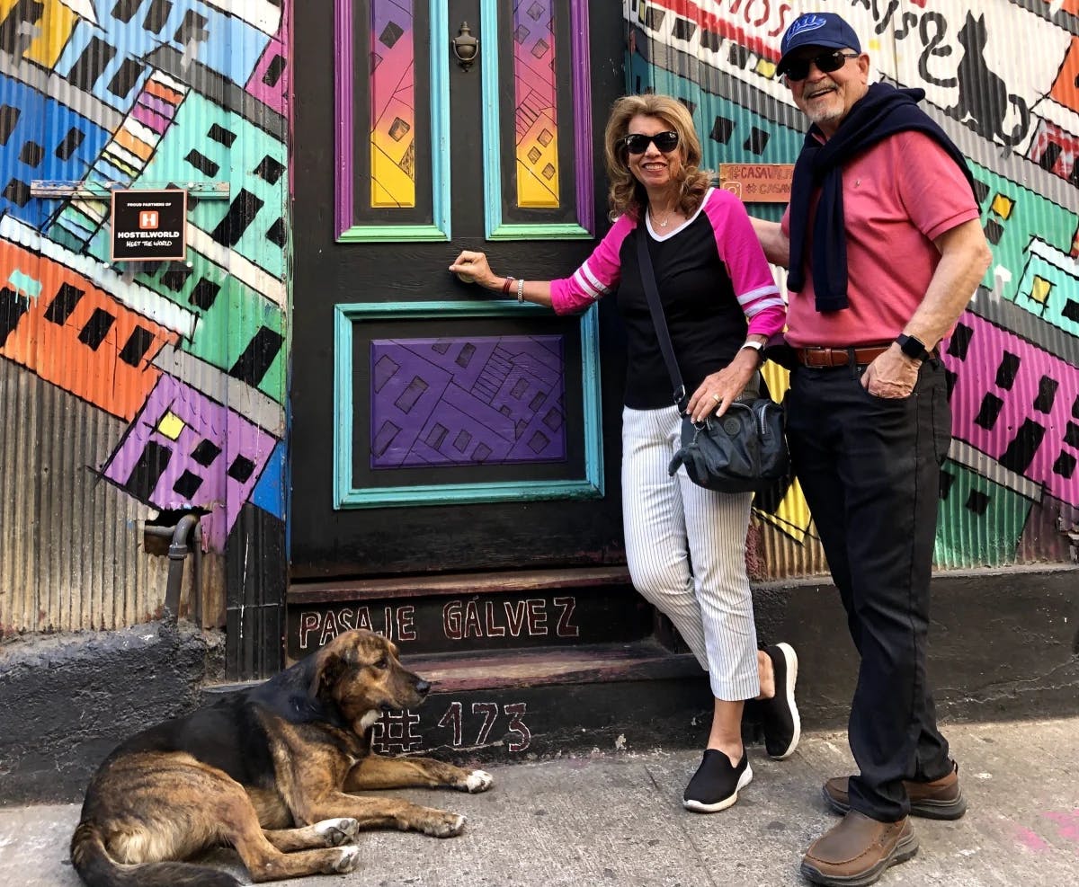 A man and woman standing in front of a colorful door and mural with a dog lying by their side.