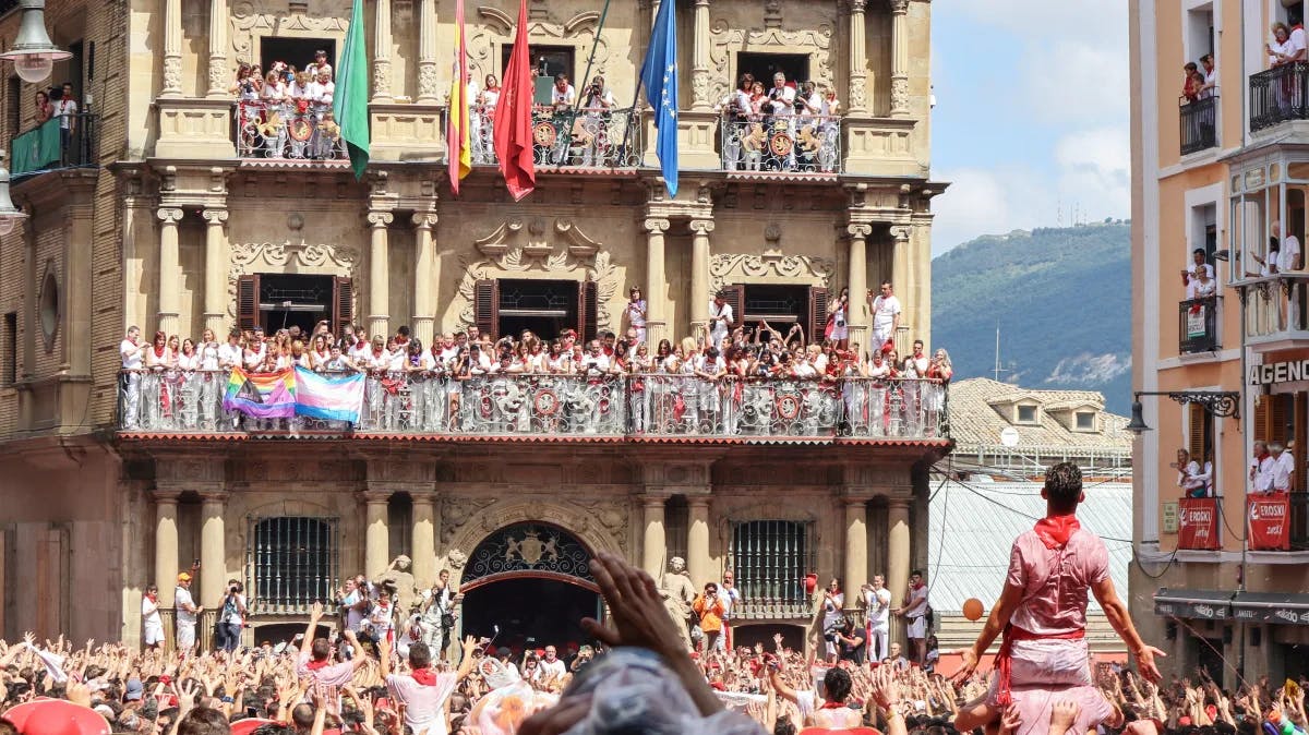 A picture of a group of people outside cheering with colorful flags while standing on a balcony against a beautiful stone building.