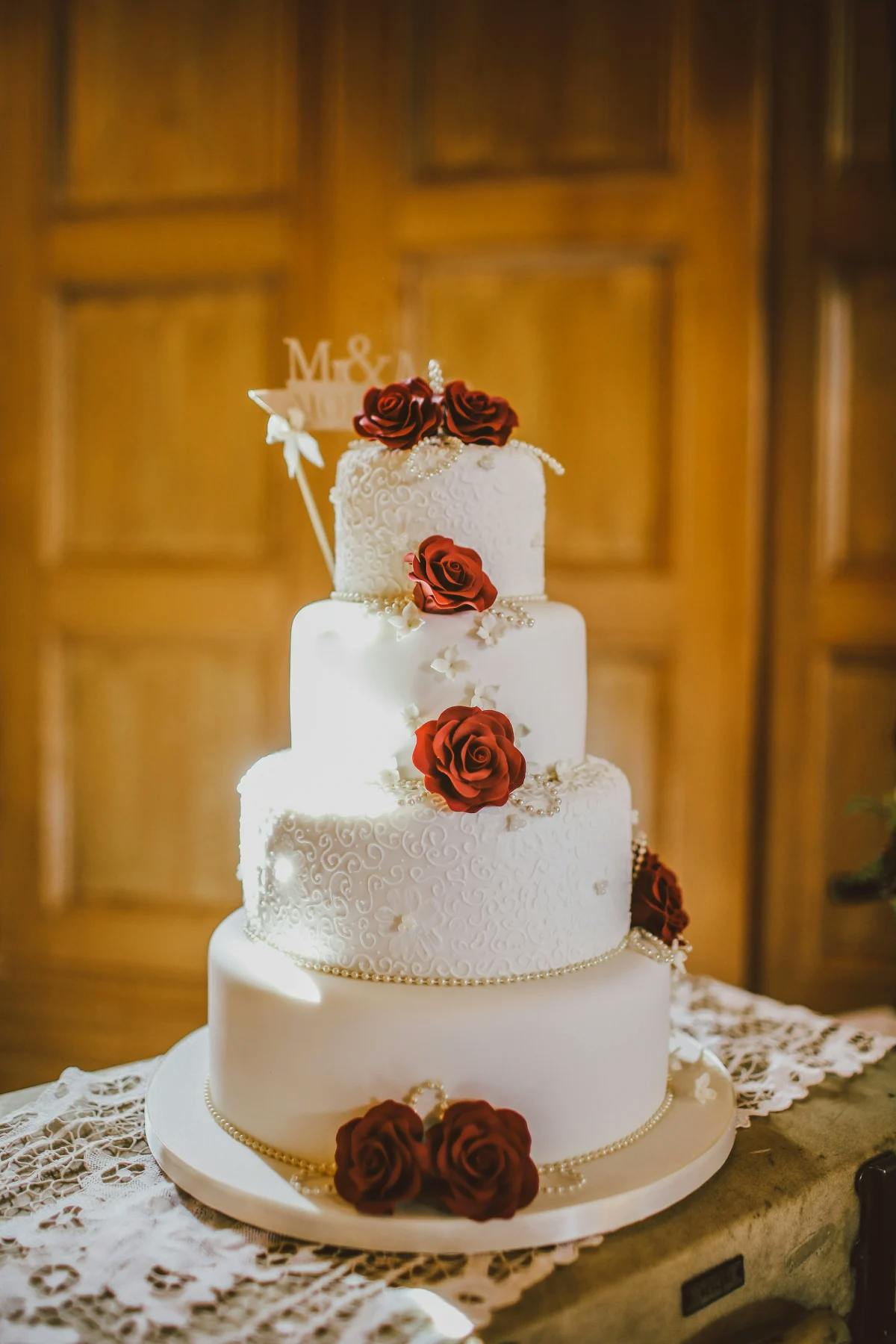 A white, four-tiered wedding cake, decorated with red roses on a wooden table with a wooden door in the background.