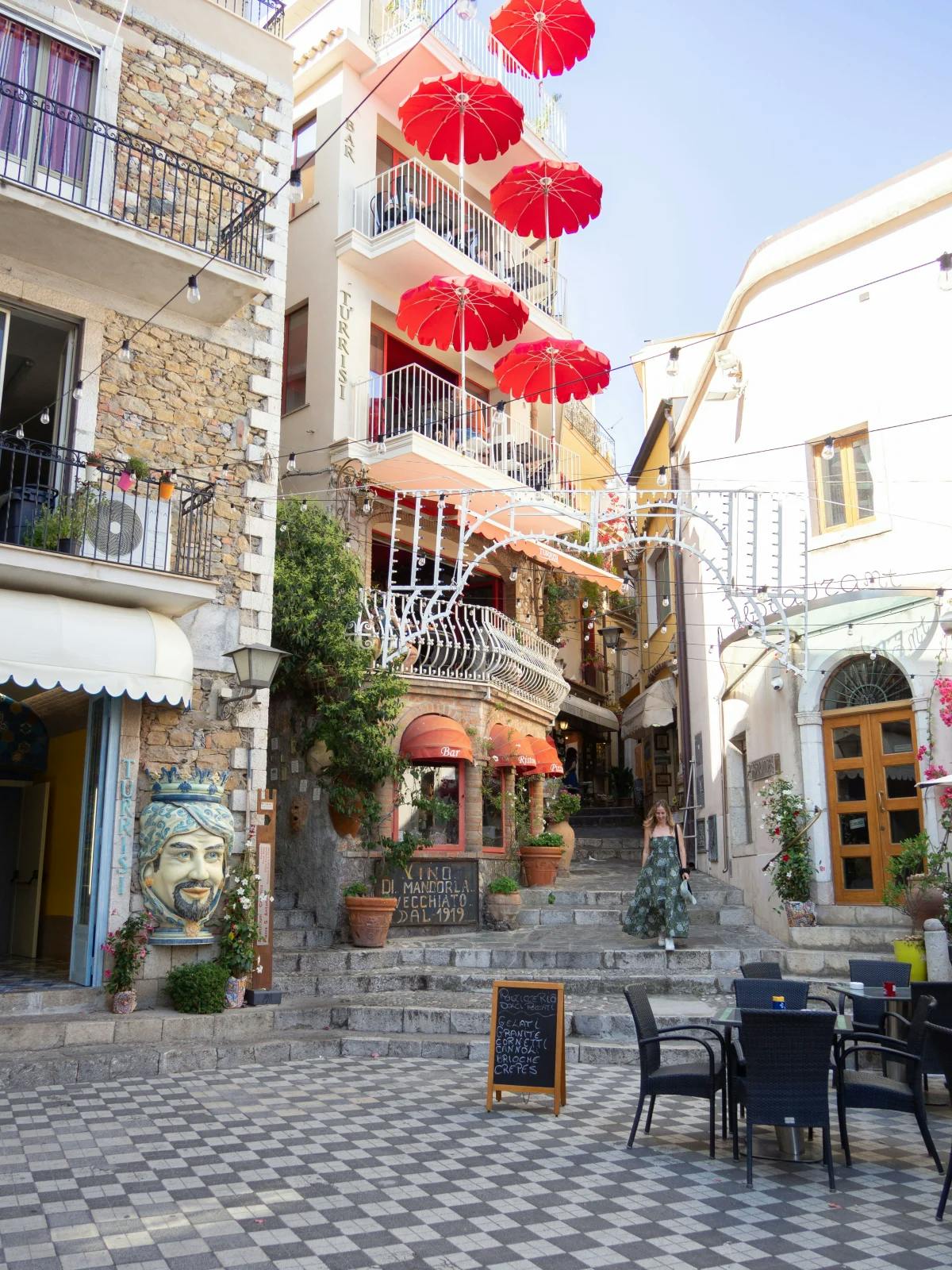 A Sicilian street scene of a local outdoor café, a woman descending down a stone staircase and residential buildings and balconies with festive decorations on a sunny day.