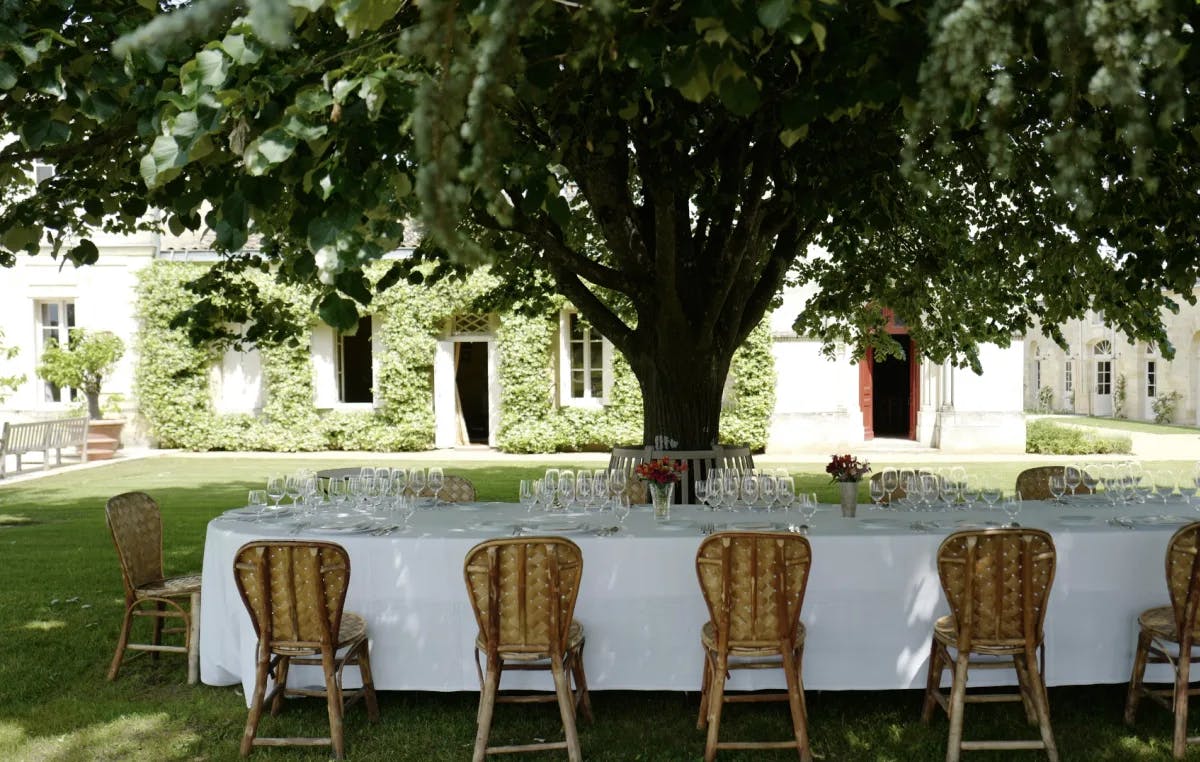 A beautiful outdoor dining table with wicker chairs, a manicured lawn, abundant tree and stone house covered in vines in the background. 