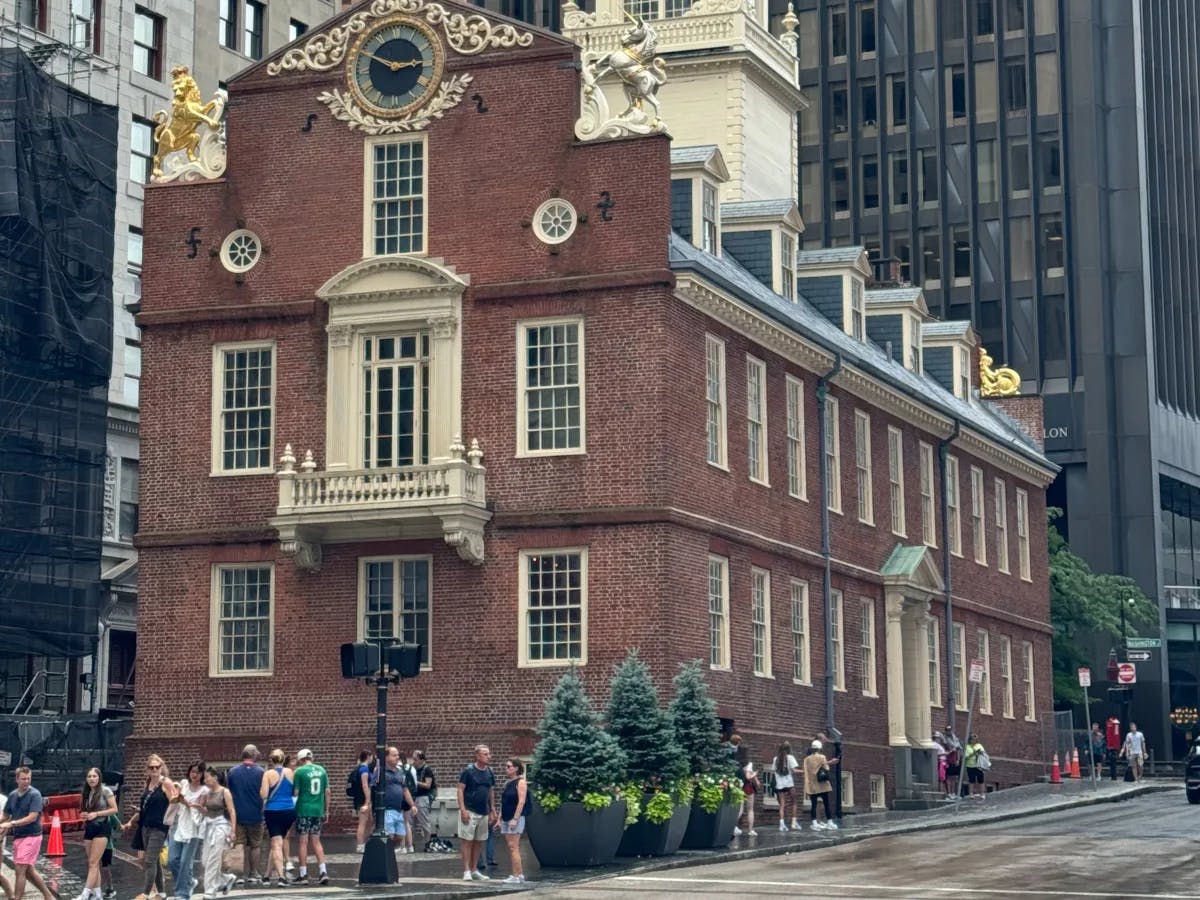 The image shows the Old State House in Boston, a historic red brick building with a white balcony, surrounded by modern skyscrapers.