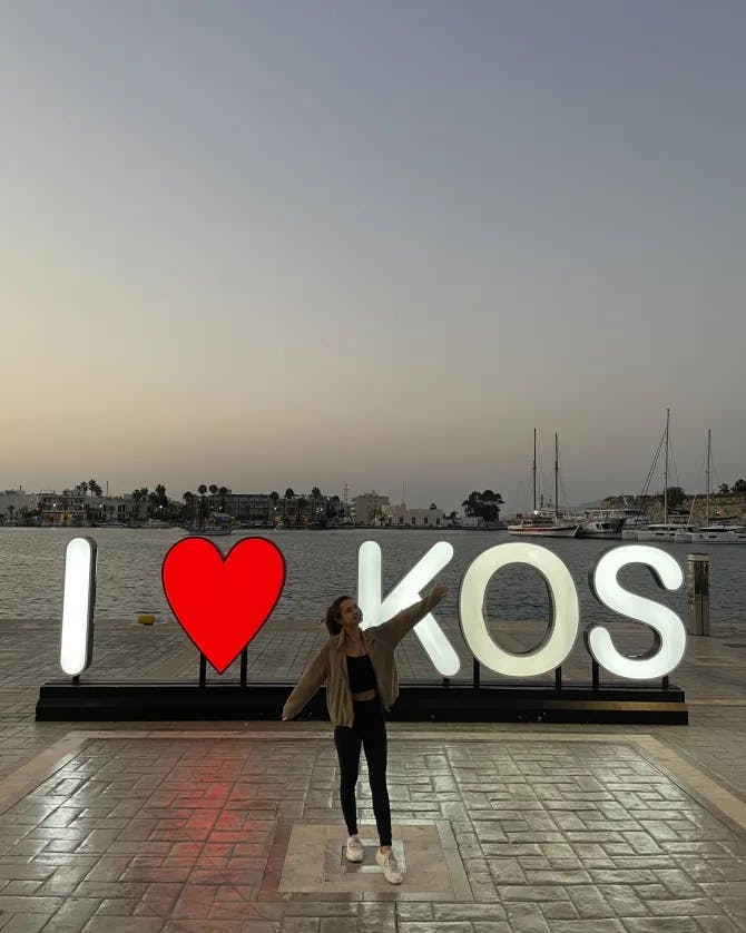 Alayna standing in front of a lit up sign reading “I <3 KOS” at sunset