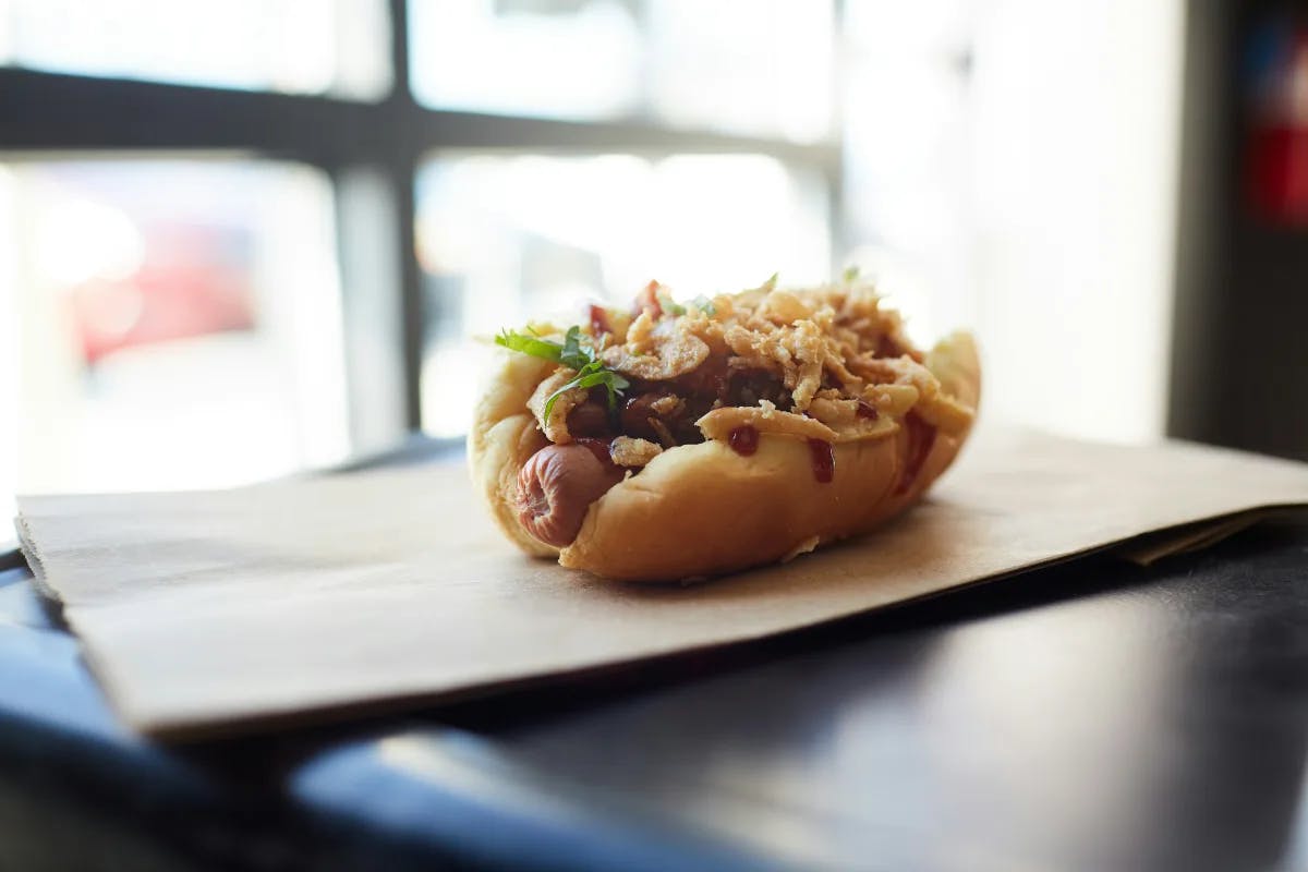 A gourmet hot dog with fried onions on a paper bag in front of a window.