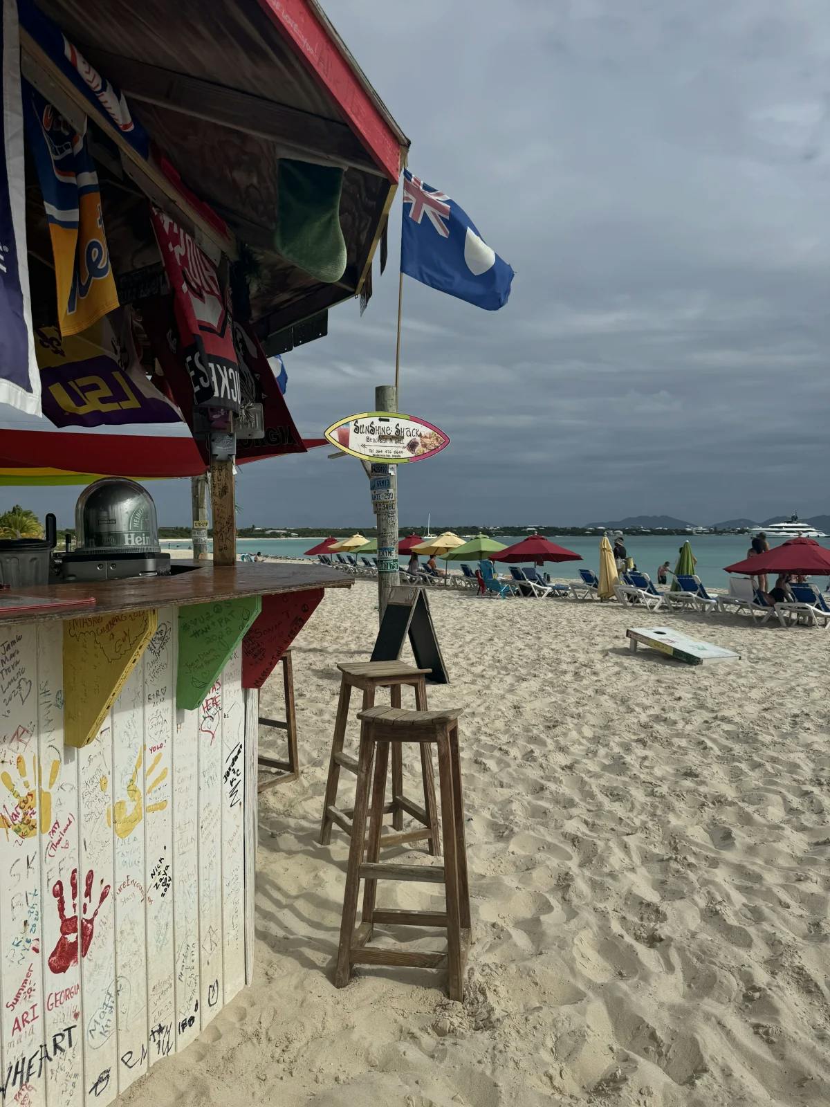 A view of a colorful beachside shack on a white sandy beach with people  on sun loungers and a flag flying